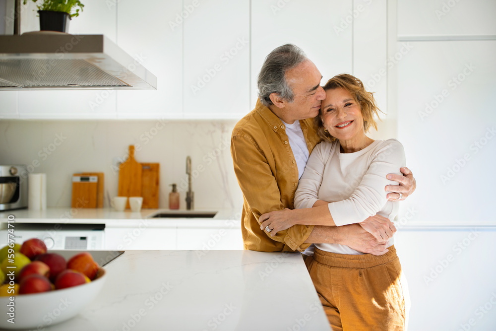 Happy loving senior spouses in casual outfits embracing, enjoying time together at cozy kitchen, man kissing his wife