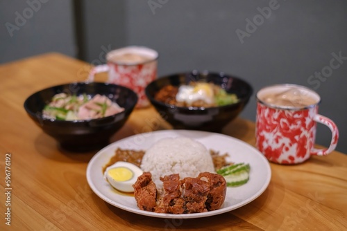 A set of Malaysian cuisine contained Steamed Chicken Rice, Nasi Lemak, Chili Pan Mee, and two mugs of Thai Tea on a wooden table with blurred background