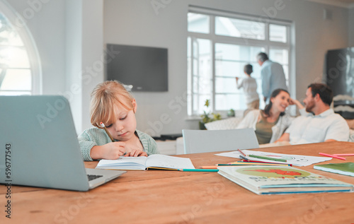 Serious young little girl writing in a notebook doing her home work while sitting at a table in the lounge at home. Small caucasian child focused on drawing in a book while using a laptop