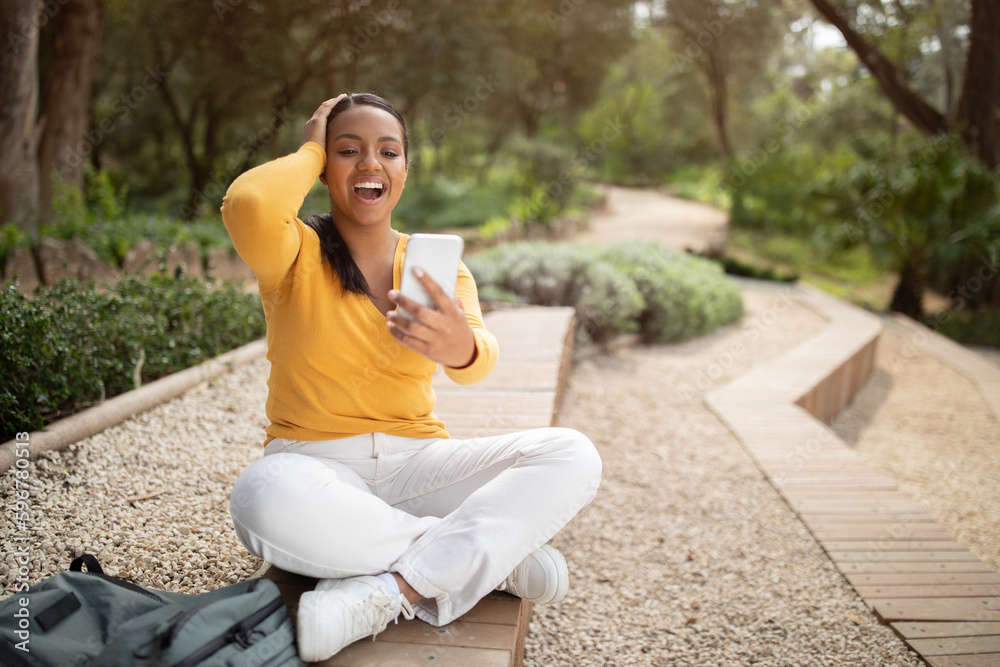 Wow. Excited young brazilian lady reading great news on smartphone and touching head, sitting in park outdoors Wow. Excited young brazilian lady reading great news on smartphone and touching head, sitting in park outdoors