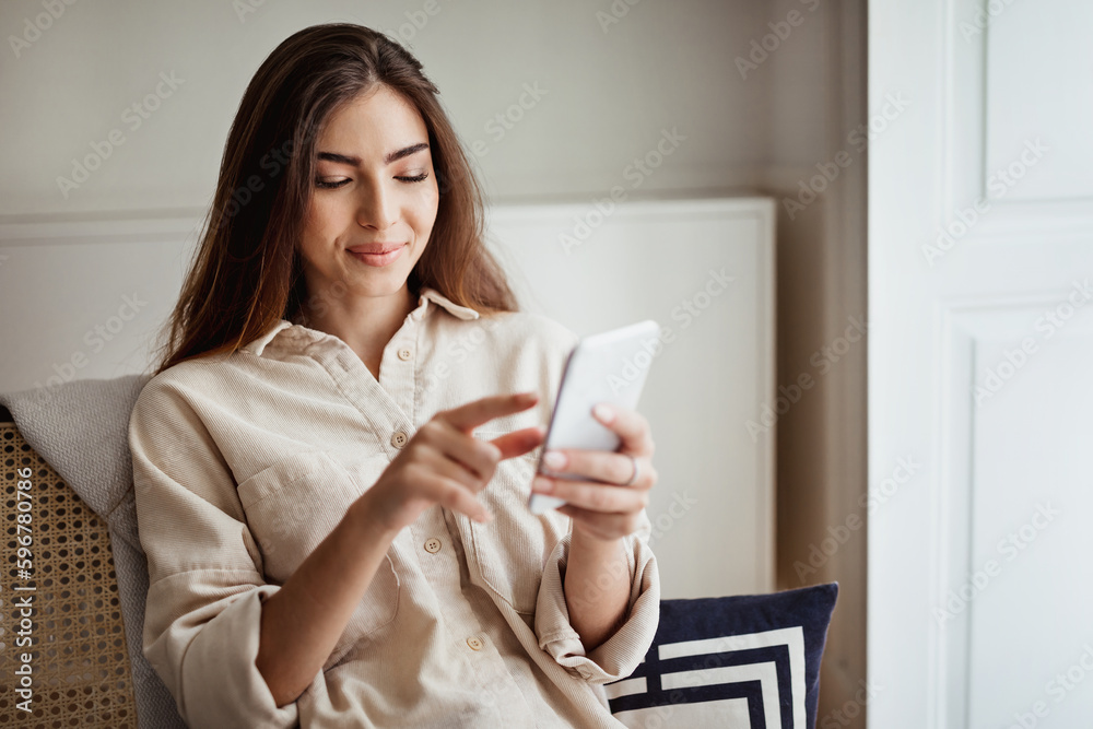 Cheerful millennial european female typing on phone, surfing in internet in minimalist living room Cheerful millennial european female typing on phone, surfing in internet in minimalist living room