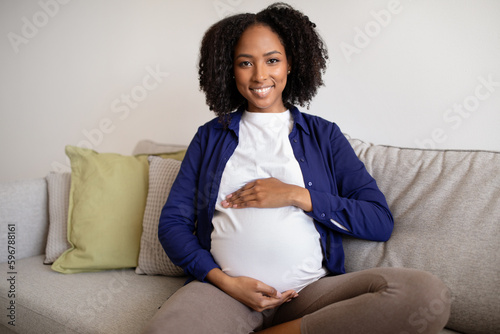 Smiling young african american curly pregnant lady in casual sits on sofa touches big belly with hands