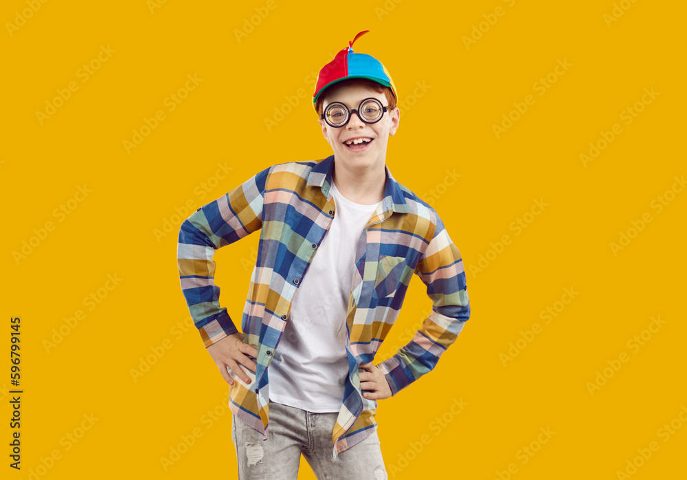 Fototapeta premium Portrait of happy, funny child in propeller cap. Cheerful, smiling little boy in casual shirt, blue red helicopter hat and round eyeglasses standing with hands on hips isolated on orange background