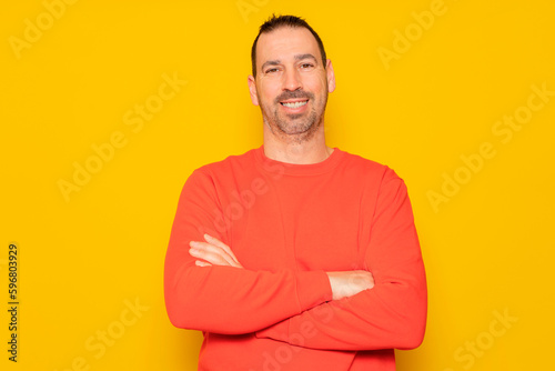 Portrait of a smart, cheerful and attractive bearded Hispanic man in his 40s, arms crossed, isolated over yellow colored background.