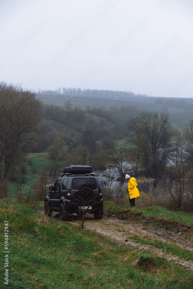 A woman in a yellow jacket next to an off-road vehicle. Overcast cold spring day, landscape with river and rocky shores. A car for tourism and extreme adventures. Vertical photo A woman in a yellow jacket next to an off-road vehicle. Overcast cold spring day, landscape with river and rocky shores. A car for tourism and extreme adventures. Vertical photo