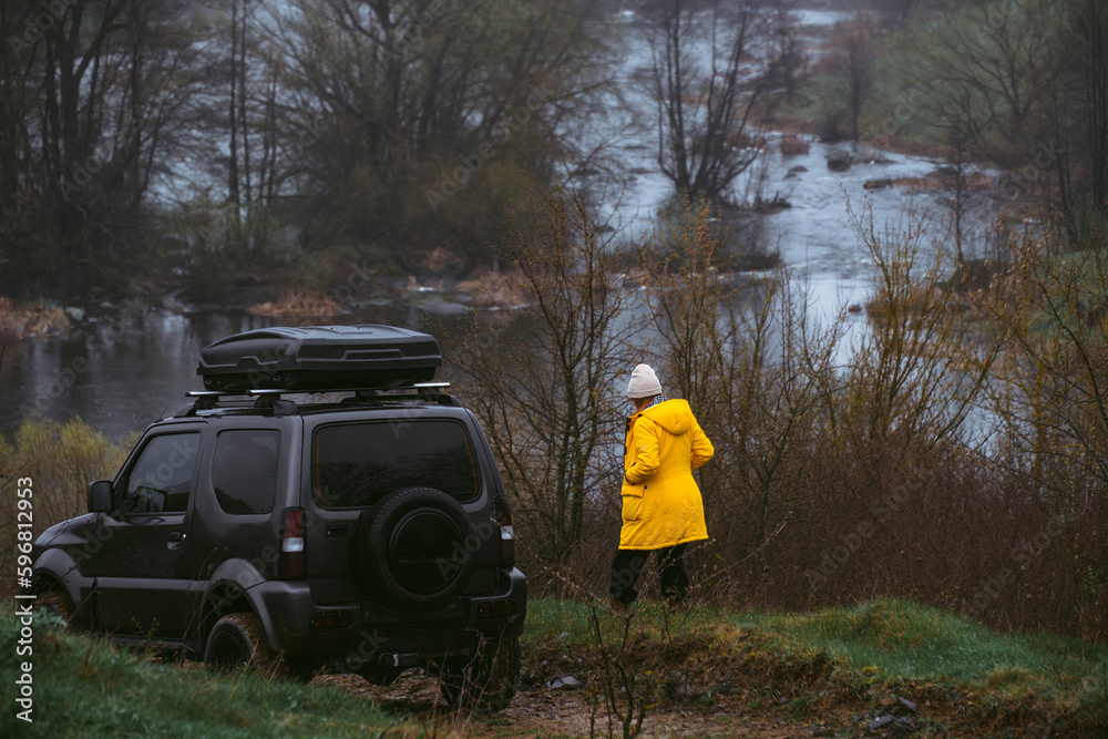 Back view a woman in a yellow jacket next to an off-road vehicle. Overcast cold spring day, landscape with river and rocky shores. A car for tourism and extreme adventures. Back view a woman in a yellow jacket next to an off-road vehicle. Overcast cold spring day, landscape with river and rocky shores. A car for tourism and extreme adventures.