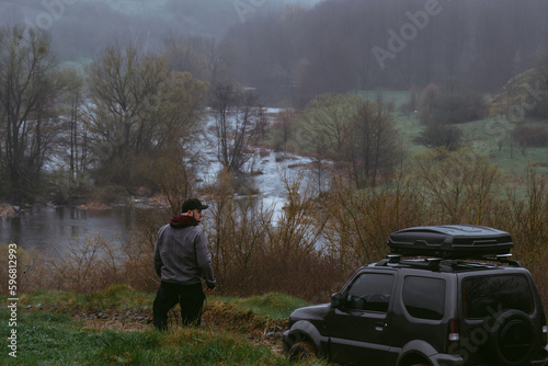 A man on top of the hill with his off-road vehicle, back view. A small compact car with four-wheel drive for extreme hobbies and recreation in wilderness. River and dirt road. Gloomy rainy spring day.