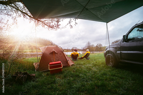 Sunbeam sun shine, a couple is sitting on folding camping chairs and having dinner with a view of the river. Breakfast red thermal container for food. Equipment for camping. Tent and off road vehicle.