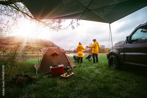 Sunbeam, couple, man and woman on vacation in wilderness. preparing the camp, The couple is celebrating. They clink beer cans.. Yellow raincoats from bad weather. A gloomy spring day. Camping by car.