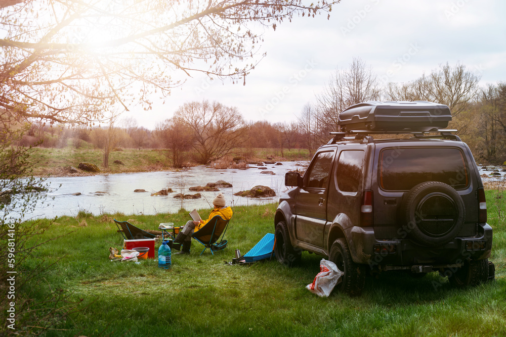 Picnic in the wilderness. An off-road vehicle with a roof rack. active recreation. Camping equipment. A woman is reading a book in nature, sitting on a folding chair, overlooking the river