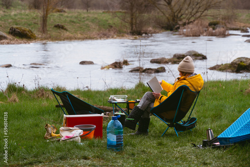 A woman is reading a book in nature, sitting in a warm jacket and hat on a folding chair, overlooking the river. Camping tourist equipment