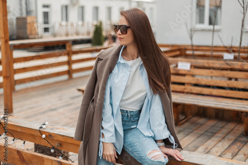 Beautiful fashionable girl with vintage sunglasses in a trendy fashion casual outfit with a stylish coat, shirt and jeans sits and rest on a wooden fence on the street