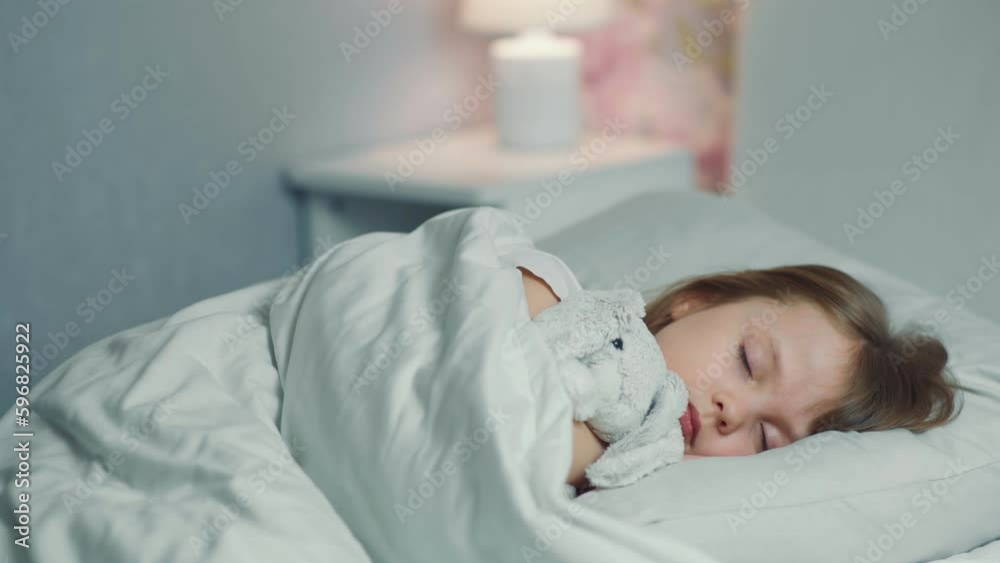 Cute funny little girl with brown hair sleeping in the white bed with favorite fluffy toy bunny. Woman hand covers with blanket caressing lying daughter. Good night. Bedtime rest. Carefree childhood Cute funny little girl with brown hair sleeping in the white bed with favorite fluffy toy bunny. Woman hand covers with blanket caressing lying daughter. Good night. Bedtime rest. Carefree childhood