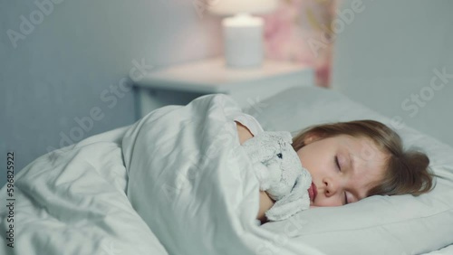 Cute funny little girl with brown hair sleeping in the white bed with favorite fluffy toy bunny. Woman hand covers with blanket caressing lying daughter. Good night. Bedtime rest. Carefree childhood