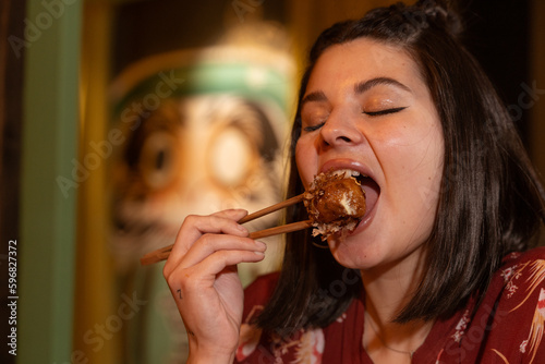 Cute brunette girl is having dinner with her boyfriend in a Japanese restaurant. She is dressed in Japanese print clothes and eats an octopus dumpling with her chopsticks.