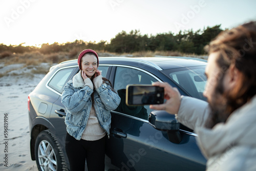 Young adult man taking a picture of a young adult woman next to a car on a beach during winter