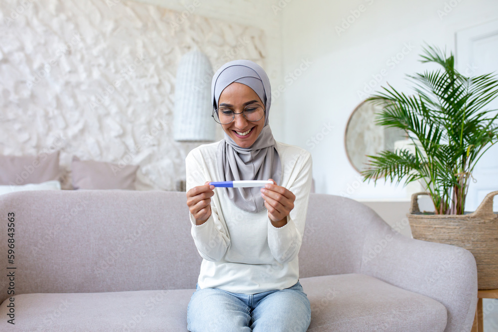 Smiling young Muslim woman in hijab sitting on sofa at home and holding ...