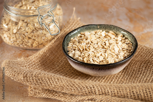 Oat flakes, cereals in a handmade ceramic bowl on burlap on a beige stone table, a glass jar with oatmeal
