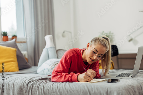 A cute caucasian college student is doing her homework on the bed.