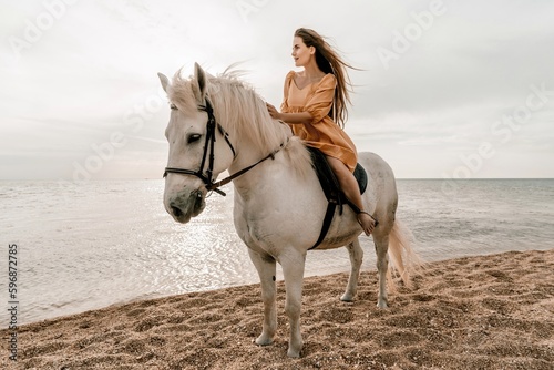 A woman in a dress stands next to a white horse on a beach, with the blue sky and sea in the background.