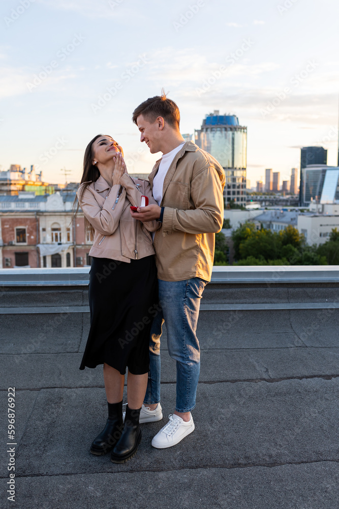 Happy young beautiful couple on a rooftop just after engagement, marriage proposal showing a ring with a rock on girl hand. Romantic date on Saint Valentine's Day. Urban cityscape on background
