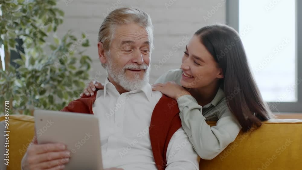 Positive senior man web surfing on digital tablet online, reading news, pretty granddaughter coming and embracing him Positive senior man web surfing on digital tablet online, reading news, pretty granddaughter coming and embracing him