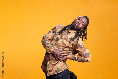 African american person suffering from backache, having lower back pain while posing in studio over yellow background. Young adult with rheumatism touching painful muscle inflammation.