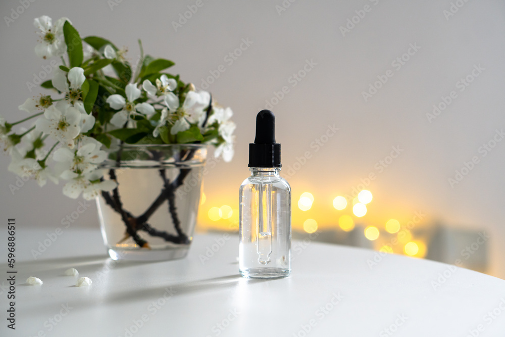 Clear glass cosmetic dropper bottle and white cherry flowers with bokeh lights, care product mockup, faded colors Clear glass cosmetic dropper bottle and white cherry flowers with bokeh lights, care product mockup, faded colors