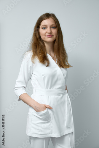 Vertical portrait of caucasian brunette female doctor wearing white lab coat smiling looking at camera and standing with her hands in pockets isolated over white wall. 