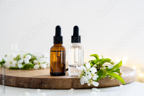 Two clear and amber glass cosmetic dropper bottles and white cherry flowers on wooden board, care products mockup