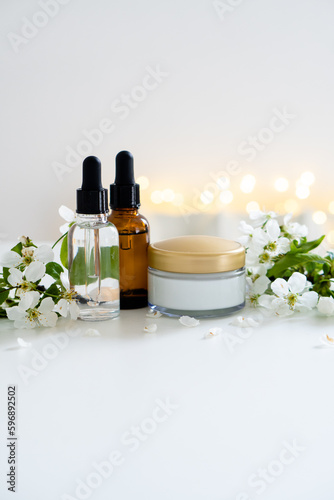 Beauty product dropper bottle and cosmetic cream jar with white cherry flowers on white table background, serum container mockup