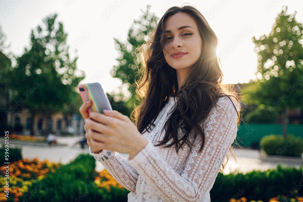 Naklejka premium Portrait of a young fashionable boho woman standing in a city park outside and using phone while looking at the camera.