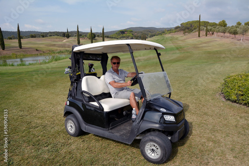 A man sits in an electric golf cart and smiles. Behind it is a large golf course