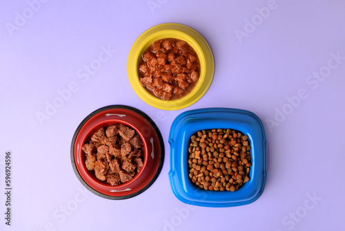 Wet and dry pet food in feeding bowls on violet background, flat lay