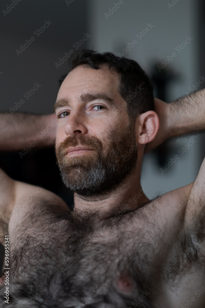 Fototapeta premium Leaning back shirtless on the sofa, a handsome man expresses assertiveness, strength, and confidence. His thick beard, chest hair, and underarm hair are part of his look.
