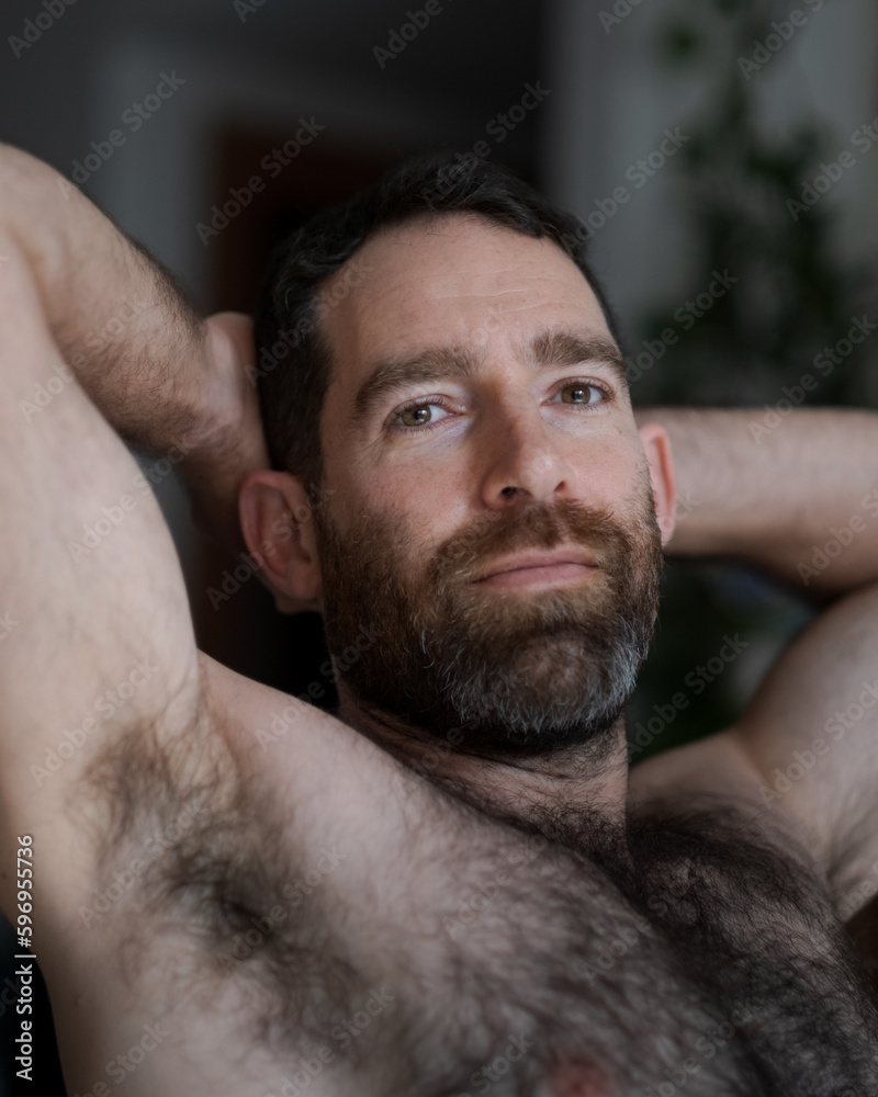 Fototapeta premium Leaning back shirtless on the sofa, a handsome man expresses contentedness, confidence, and a gentle smile. His thick beard, chest hair, and underarm hair are part of his look.