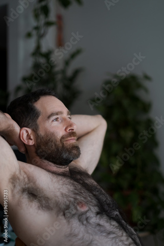 Leaning back shirtless on the sofa, a handsome man expresses being at home, comfortable and confident. His thick beard, chest hair, and underarm hair are part of his look.