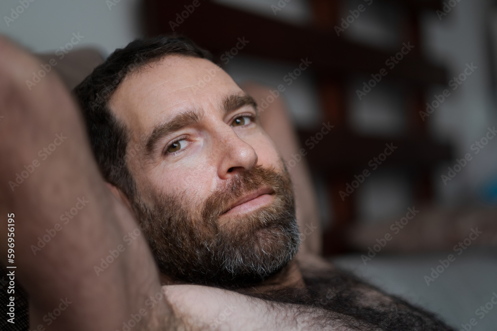 Fototapeta premium Leaning back shirtless on the sofa, a handsome man expresses a domestic moment, looking over at the camera while relaxing at home. His thick beard, chest hair, and underarm hair are part of his look.