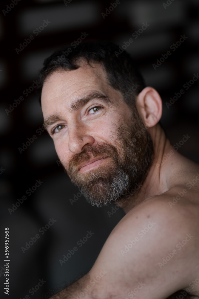 Fototapeta premium Looking over his shoulder while relaxing at home, a man expresses laughter and smiling. He is shirtless, with a thick beard that draws attention to his face.