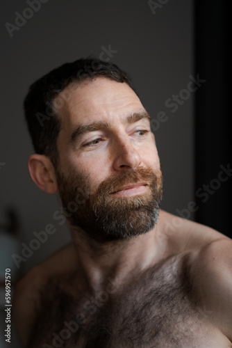 Looking over his shoulder while relaxing at home, a man expresses reflection and contemplation. He is shirtless, with a thick beard that draws attention to his face.