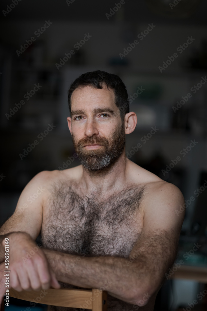 Sitting backwards on a chair, a man expresses an austere pose and neutral expression. He is shirtless, with his thick beard and chest hair drawing attention to his masculine physique. Sitting backwards on a chair, a man expresses an austere pose and neutral expression. He is shirtless, with his thick beard and chest hair drawing attention to his masculine physique.