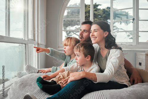 Young caucasian couple enjoying the view from a window while bonding with their adorable little children on a comfy seat. Cute kid looking amazed and pointing in surprise while sitting with her family photo