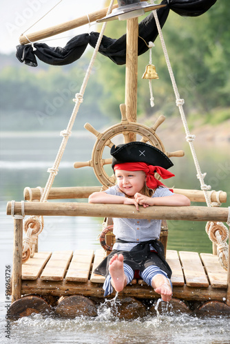 Child in a pirate costume plays on a wooden raft at sunset. Girl pretends to be the captain of a ship with black sails and a flag. Funny kid dreams of adventure and travel.