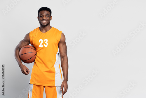 Portrait of confident smiling African American man, professional basketball player holding ball isolated on white background. Sportsman wearing orange basketball uniform looking at camera, copy space