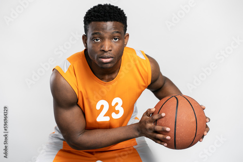 Portrait of young serious basketball player holding ball isolated on white background. Handsome African man wearing orange sportswear playing basketball looking at camera. Sport competition concept