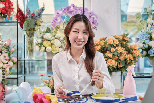 One Young beautiful Asian female florist entrepreneur arranging a bunch of blossoms, decorating with lovely ribbons, happy work in colorful flower shop store with blooms, and small business owner.