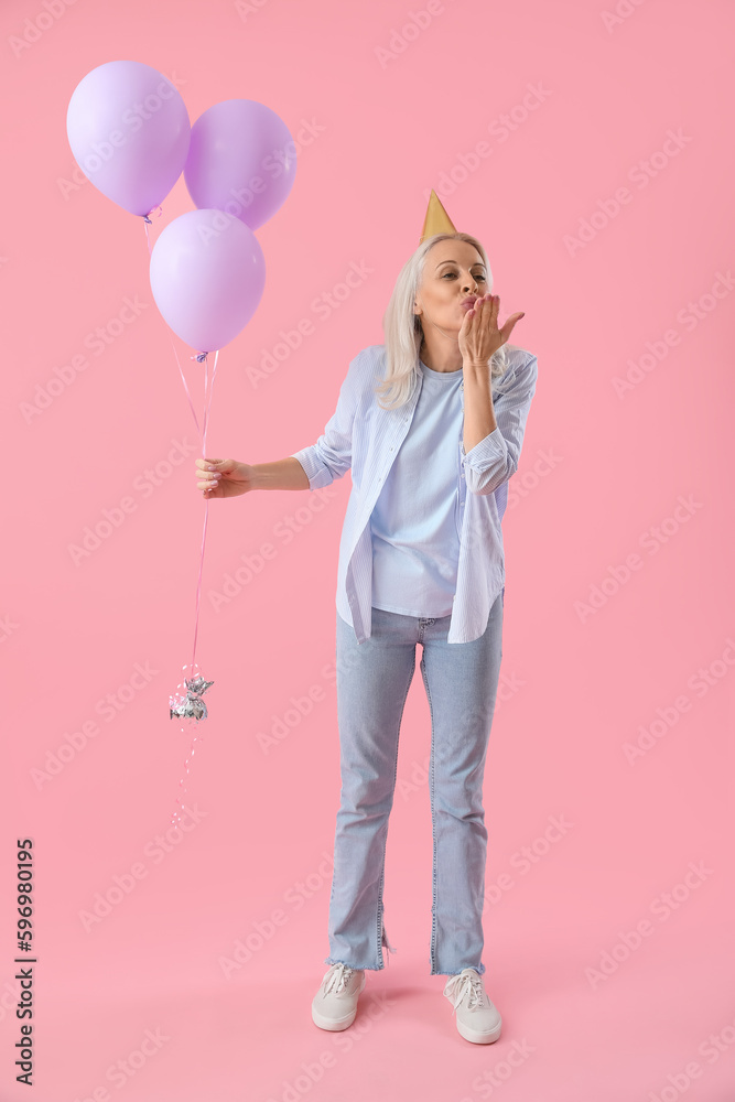 Mature woman in party hat with balloons blowing kiss on pink background Mature woman in party hat with balloons blowing kiss on pink background