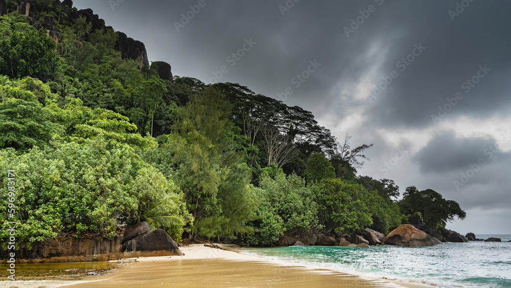 Rainy day on a tropical island. The waves of the turquoise ocean spread over the wet sand of the beach. Lush tropical vegetation on the hill, boulders in the water. Cloudy. Seychelles. Mahe. Rainy day on a tropical island. The waves of the turquoise ocean spread over the wet sand of the beach. Lush tropical vegetation on the hill, boulders in the water. Cloudy. Seychelles. Mahe.