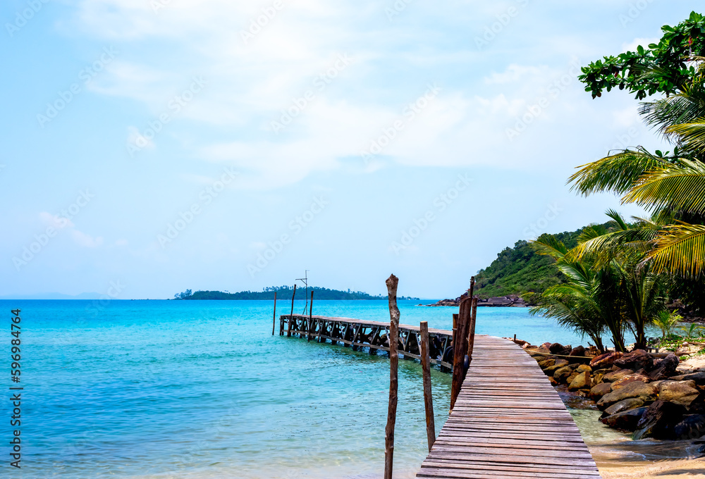 Wooden bridge heading to the blue sea. Brown wood plank pathway bridge on the beach sand with rocks and coconut palm tree at the local port in island on sunny day. Seascape summer holiday background.