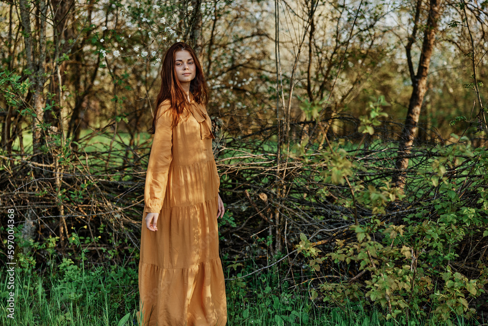 a beautiful woman walks in the shade near the trees, dressed in a long orange dress, enjoying the weather and the weekend, posing relaxed. The theme of privacy with nature a beautiful woman walks in the shade near the trees, dressed in a long orange dress, enjoying the weather and the weekend, posing relaxed. The theme of privacy with nature